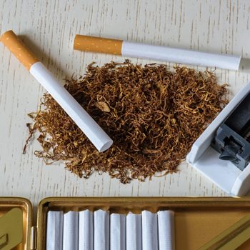 A pile of natural tobacco on a white wooden table, a device for manual production of cigarettes and cigarettes in a cigarette case. Smoking. Nicotine addiction. Health hazard.