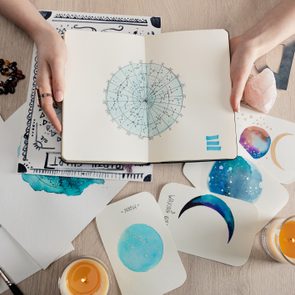 Top view of astrologer holding notebook with watercolor drawings and zodiac signs on cards on table