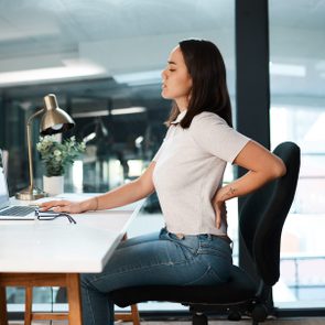 young woman suffering from back pain while sitting and working at desk in office