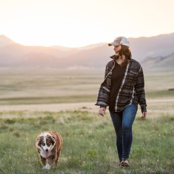 woman walking dog in a field with mountains in the background during sunset